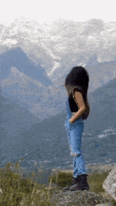 a woman in overalls is standing on a rock looking at a mountain range .