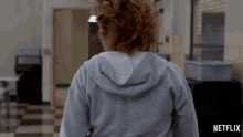 a woman in a grey hoodie is walking down a hallway with a netflix logo on the bottom right