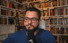 a man with glasses and a beard is sitting in front of a microphone in front of a bookshelf .