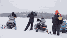 a person laying on a snowmobile while another person stands nearby