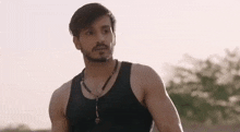 a man with a beard wearing a black tank top and a necklace is standing in the desert .