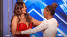 a woman in a red dress is hugging another woman in a white dress with the nbc logo behind them