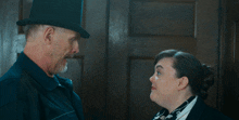 a man in a top hat talks to a woman in a black jacket