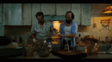 two men in a kitchen with a christmas stump poster on the wall