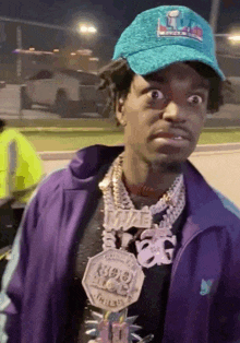 a man wearing a purple jacket and a blue hat has a necklace that says ' wayne ' on it