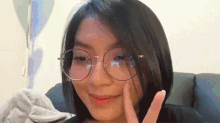 a girl wearing glasses is sitting on a couch giving a peace sign .