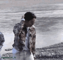 a man in a camouflage jacket is standing on a rocky beach near a body of water ..