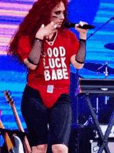 a woman singing into a microphone wearing a red shirt that says " good luck babe "