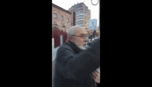 an elderly man with a beard and glasses is talking on a cell phone on a city street .