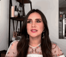 a woman with long dark hair is wearing a floral top and earrings and looking at the camera .