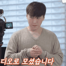 a man in a tan sweater stands in front of a camera with korean writing on his arm