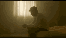 a man sits on a bed in front of a window with his hands folded