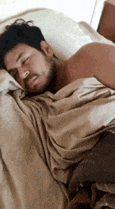 a man with a beard is sleeping in a bed with a brown blanket .