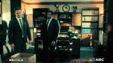 two men in suits are standing in a room with nbc on the bottom