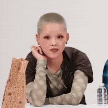 a woman with a shaved head sits at a table next to a paper bag