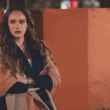 a woman in a coat is standing with her arms crossed in front of a wall .
