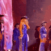a woman in a blue jumpsuit and cowboy hat stands on a stage