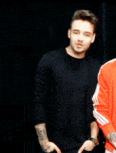 a man with a tattoo on his arm is wearing a black shirt and a red jacket .