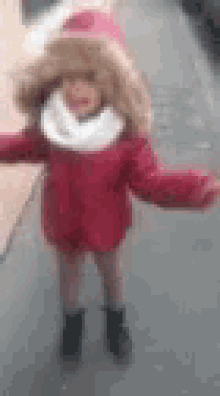a little girl wearing a red coat and a fur hood is standing on the sidewalk .