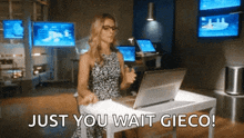 a woman is sitting at a desk with a laptop and says just you wait gieco !