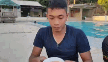 a young man in a blue shirt is sitting at a table with a plate of food in front of a swimming pool .