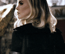 a woman with blonde hair and a black jacket looks over her shoulder