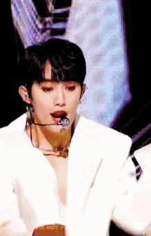 a man in a white jacket is singing into a microphone while wearing a necklace and earrings .