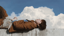 a man in a brown jacket is laying on a wall with a cloud in the background