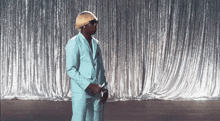 a man in a light blue suit and wig is standing in front of a silver curtain .