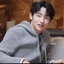 a young man in a gray sweater is sitting in front of a chalkboard with mathematical equations on it .