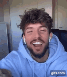 a man with a beard wearing a blue hoodie and ear buds is smiling .