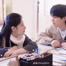 a boy and a girl are sitting at a table looking at each other with chicophim written on the bottom right