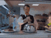 a man in a tuxedo is holding a glass dome in a restaurant kitchen .