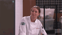 a woman in a chef 's jacket is sitting in a kitchen with bravo in the background .