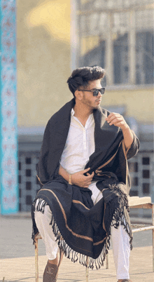 a young man wearing sunglasses and a black shawl sits on a chair