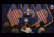 a man stands at a podium in front of american flags with cnbc live on the screen behind him