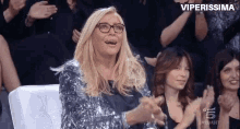 a woman wearing glasses and a sequined dress is sitting in a crowd of people applauding .