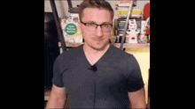 a man wearing glasses and a gray shirt is standing in front of a shelf .