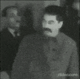 a black and white photo of a man with a mustache standing in front of a group of men .