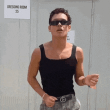 a young man wearing sunglasses and a black tank top is standing in front of a sign that says dressing room 25 .