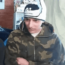 a man wearing a camouflage hoodie and a beanie is sitting in a room .