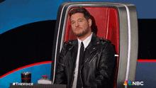 a man in a leather jacket and tie sits in a chair with a nbc logo on the bottom