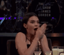 a woman is sitting at a table with a martini in front of a sign that says the late show with james corden .