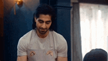a man wearing an apron and a white t-shirt is standing in front of a window .
