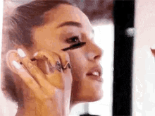 ariana grande is applying mascara to her eye while looking in the mirror .