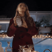 a woman in a red coat is standing in front of a swimming pool holding a glass