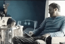 a man is sitting in a chair with a baby in a crib while using a tablet .
