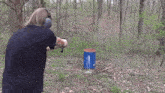 a man is shooting a gun in the woods near a blue barrel