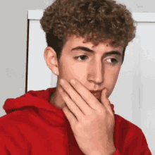 a young man with curly hair wearing a red hoodie covering his mouth with his hand