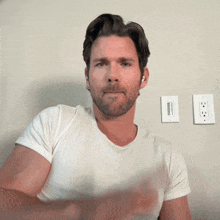 a man in a white shirt is standing in front of a wall with two electrical outlets on it
